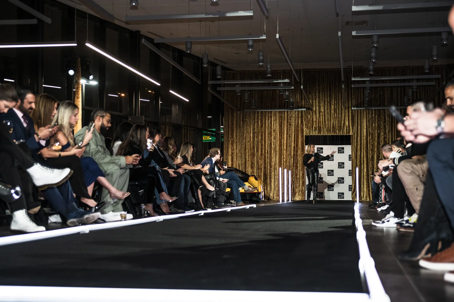 A fashion event runway with guests seated on either side and a gold curtain stage.